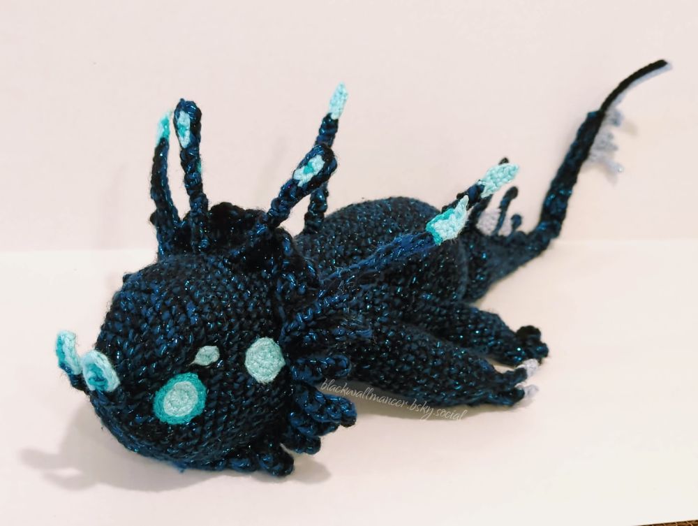 A crocheted Deep Dweller from Divinity: Original Sin 2. It's lying on its stomach with its arms out at its sides in a "sploot"-like pose. Displayed on a white backdrop, the piece is 24" long (61cm), 7" tall (18cm), and 9" wide (23cm). Crocheted with dark blue and black sparkly acrylic yarn (held double) and stuffed with Poly-Fil, it has several antennae along its head and back (with lighter blues on the tips to give it the appearance of glowing lights), along with fins (in a lighter grey/blue) atop and at the tip of its tail. The front legs end in clawed hands, while the rear legs end in fins.

Freehanding this was a complete and utter pain, but obviously there aren't patterns out there for super-specific fandom stuff like this, so I did what had to be done and tortured the fuck out of myself. Worth it, though!