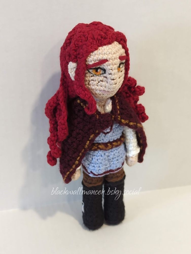 A crocheted doll of Albtraum, an OC with scarlet hair, very light skin, orange eyes, and freckles. His hair is a little curly and reaches far down his back. He's wearing black boots with silver buckles, dark brown trousers, a blue tunic with mauve embroidery around the sleeves, collar, and hem, and a white undershirt with long sleeves and a collar. His tunic is held tight to his body by a mauve belt with gold embroidery, and around his shoulders a short cloak is fastened (mauve with gold embroidery). He is about nine inches tall.