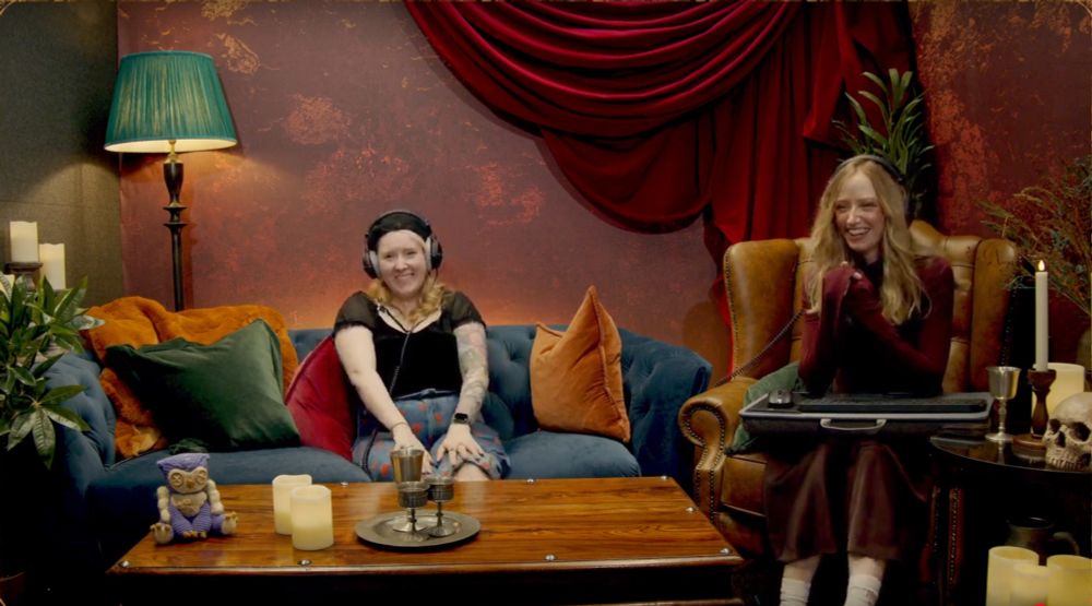 Leah (@leahlemoncakes.bsky.social) and Aoife on Larian's BG3 stream today, both smiling and talking while playing a Dark Urge run. In what looks like a cozy corner of a living room, Leah sits on a couch covered with pillows, while Aoife sits in a tall armchair. In front of them is a coffee table, atop which are a few candles, a tray with goblets(?), and one of the owlbear amigurumis I crocheted! wow, he's famous!