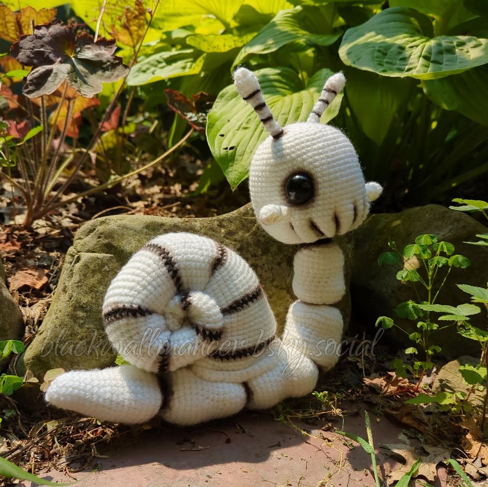 A photo of a crocheted snail skeleton sitting in a garden. The snail is crocheted with white yarn and features black accents, with the black parts enhanced with brown pigment to add depth to the appearance of the bones. The snail has a big head with large dark eyes, stalks on the top of its head and under its eyes, and five "spine" bones connecting the skull with the shell. Listen, it's kind of hard to describe a skeleton snail since snails don't have skeletons. Anyway, it's pictured here on a flat stone path in front of a small garden (also surrounded by stones). The plants are green and shades of orange and gold. Hurry up, fall!

Pattern by Cottontail & Whiskers

Snailetor is 11 inches tall (28cm) x 11 inches long (29cm). The "spine" is wired for some poseability.