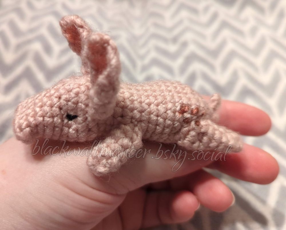 Pictured resting on my hand, mostly lying across my thumb, is a small crocheted nug (a rabbit/pig sort of creature from the Dragon Age series). It's a soft pink, with brown spots on its rear and a darker pink in its ears. It has a tiny tail that curves upward, and its snout curves down.