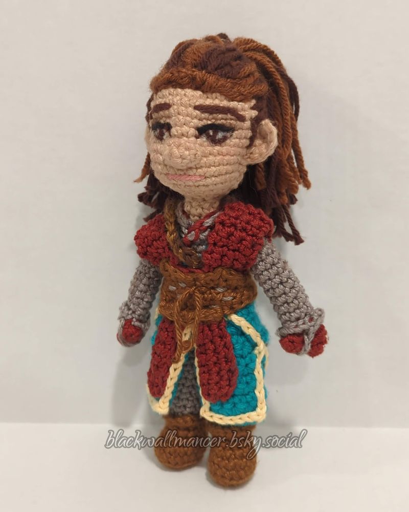 A crocheted Rook from DA:TV. She has dark brown hair with reddish-brown highlights and soft brown eyes, and is wearing turquoise and red Battle Robes with brown belts and boots. The clothing beneath her robes is grey, and it covers most of her body and her arms, except for her hands, where her red gloves peek through.