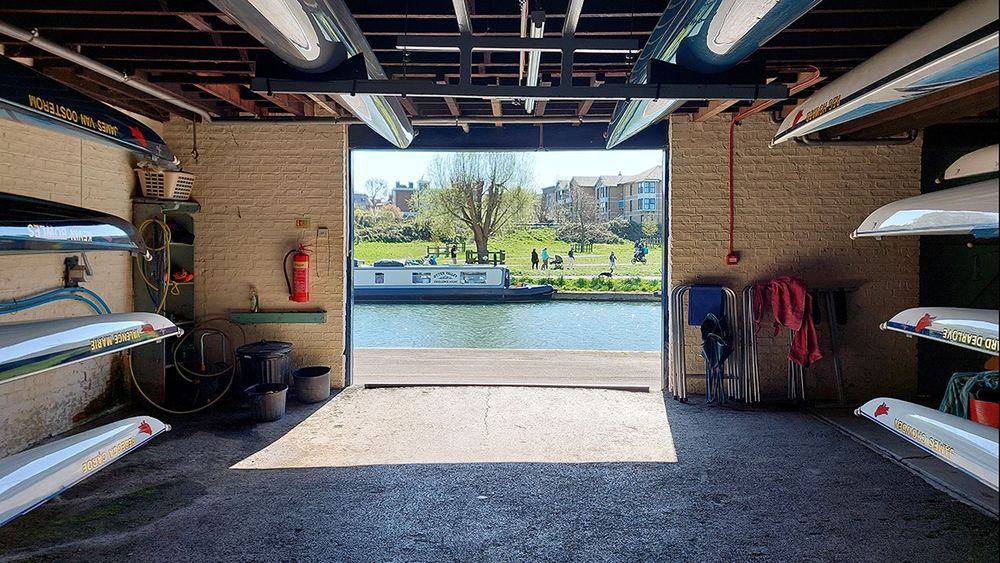 The view out of Pembroke Boat House, towards the Cam. Pembroke boats are stored in stacks on the left and right of the picture, with two suspended from the ceiling. Across the river, it's a sunny day, with people walking across Midsummer Common and a blue and white houseboat moored on the far bank.
