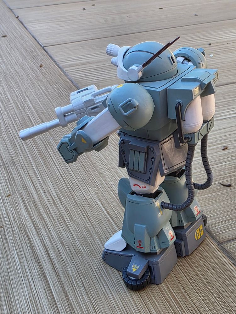 Assembled Votoms gunpla model kit, painted with Love Bullet blue/white/grey livery and holding a weapon with a heart-shaped barrel opening. 3/4 rear view.