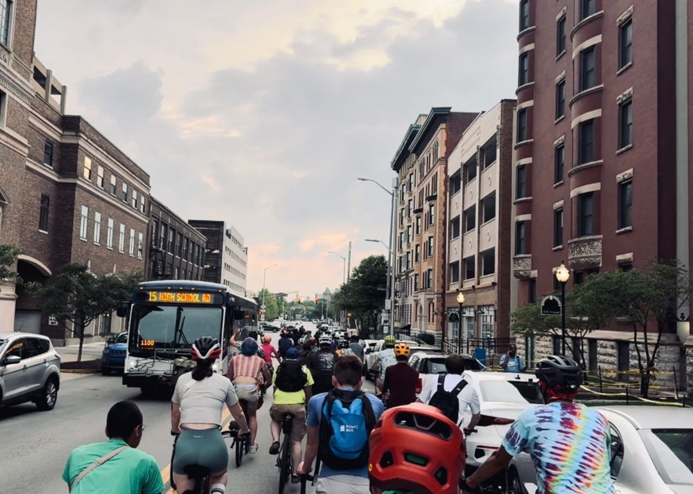 A photo of approximately 75 cyclists cycling down a street with the #15/High School Rd bus driving in the opposing direction. 