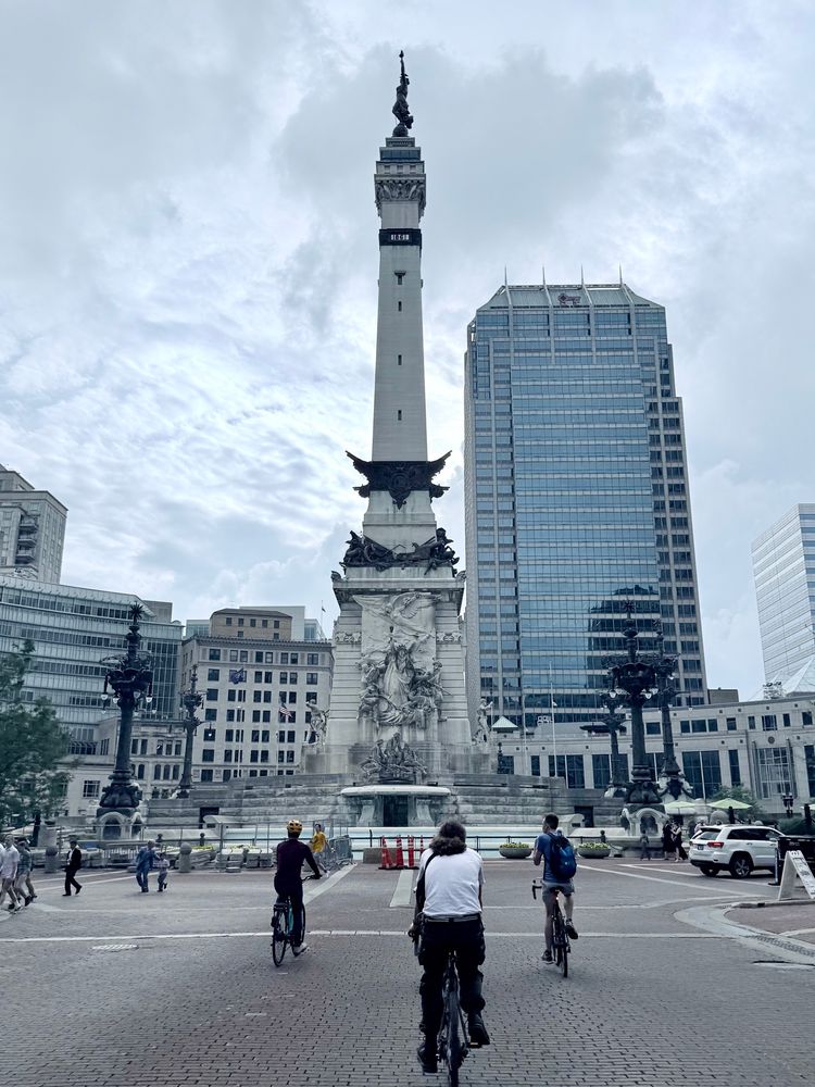 A photo of the Soldiers’ and Sailors’ Monument in Indianapolis. Numerous cyclists and pedestrians are seen navigating the traffic circle around the monument. 