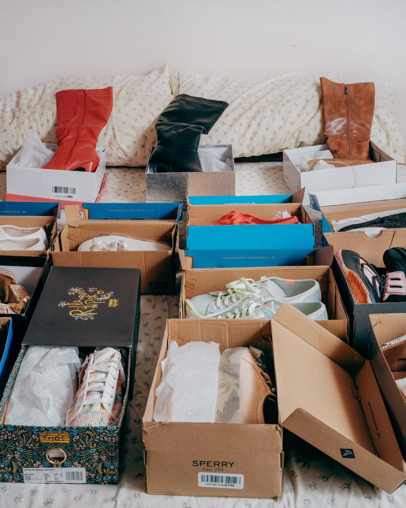 Various pairs of shoes, including boots and sneakers, are displayed in open boxes on a bed with a white blanket. 