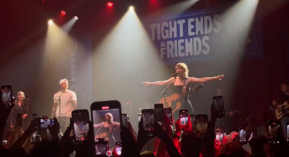 taylor swift performing shake it off at a tight end university event in nashville. she’s wearing a black dress and has a guitar on her. her arms are up and you can see the crowd looking at her and recording her with their phones.