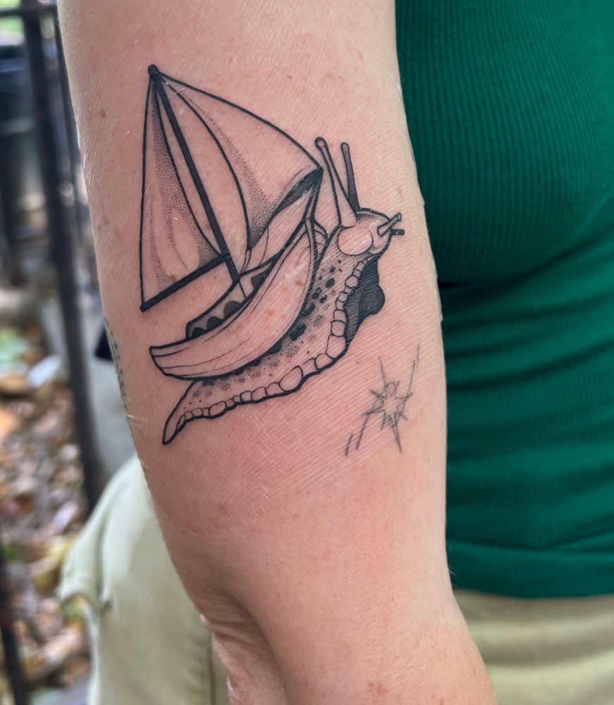 A black tattoo on a white arm of a snail/sea slug carrying a sailboat on its back through the stars. Artist is Phia Walla and they have an incredibly light hand. Very easy tattoo to sit for.