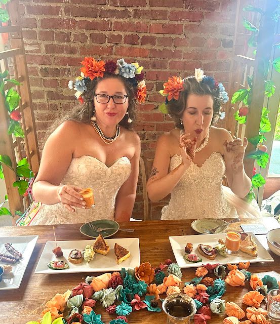 A white woman with long light brown hair and a white non-binary person with short dark hair streaked with grey. They are both wearing wedding dresses and flower crowns and are seated at a table eating plates of appetizers at their wedding. Hand-dyed wooden flower in shapes of blue, peach, and brick red decorate the table.