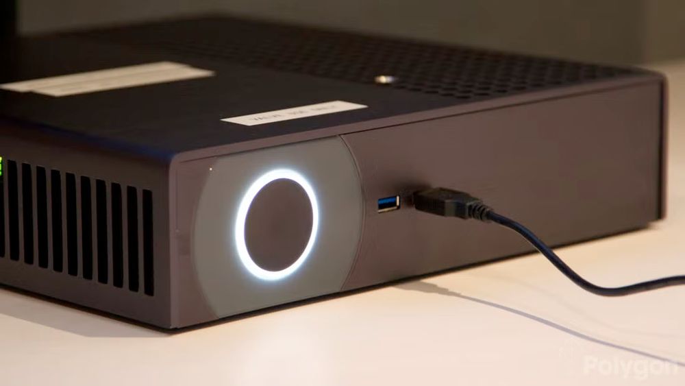 A Steam Machine prototype is pictured sitting diagonally on a surface within a room. The unit is rectangular and has a minimalist matte black design. The front panel has a LED ring around a power button, and 2 USB ports, these are situated on one end of the panel, with the rest being an empty rectangle. The side facing the camera is rounded toward the top, and has slots cut out for air flow. The top is partially a solid surface, and partially perforated with rounded holes. There appear to be labels affixed to the top panel of the unit, but it is unclear what they say or what purpose they serve.