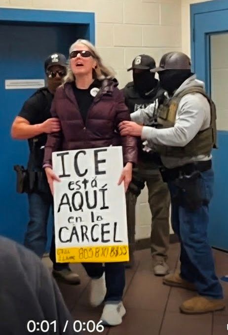 Three Ice agents grabbing and pushing a tall white lady with gray hair holding a sign warning ICE esra aqui en la Carcel. In SLO County CA jail yesterday 12/14 at 10:00 am. 