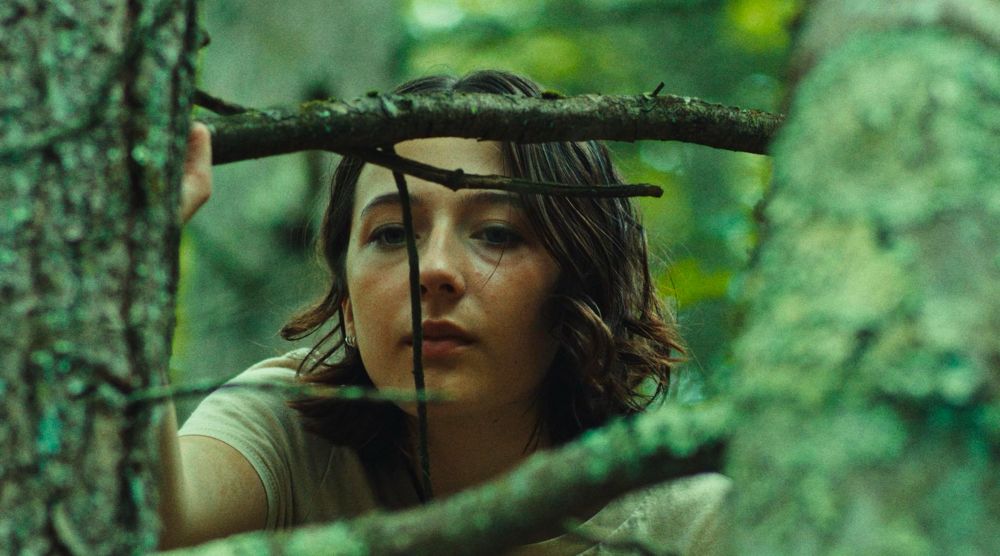 Still from Good One featuring a close-up of a young woman peering through intertwined branches in a forest