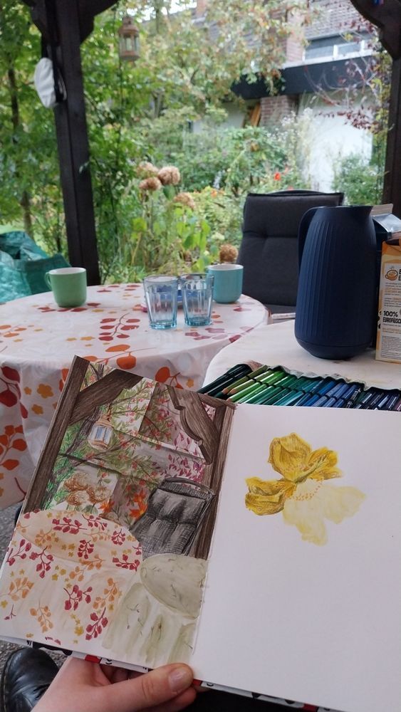Photograph of sketchbook held up in front of some tables and a garden chair with plants in the background. The tables are covered with flowery tablecloths. There's a dark grey/black folding chair positioned slightly to the right behind the tables. Two dark wooden beams behind it sort of frame the greenery in the background. A rose is growing along the left beam. There's a tree with what looks like bright red, tiny apples in the far left background. There's a tree with purple-ish, dark red leaves in the far background to the right. On the bottom there's some dried white-brownish flowers and some bright orange ones. The sketchbook shows a drawing of that same scenery on the left page. On the right page there's a drawing of a single yellow flower of Great St. John's Worth. Both drawings have a watercolour base with pencil structures and plants on top. Coffee cups and other coffee things as well as a roll of pencils spread on the table are left out of the drawing.