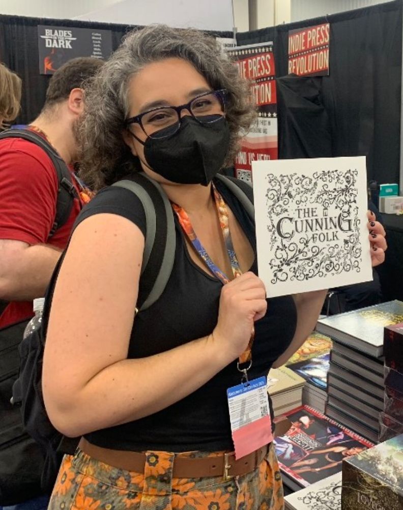 Emily looking happy and wearing a mask, holding up a copy of The Cunning Folk at the IPR booth at GenCon