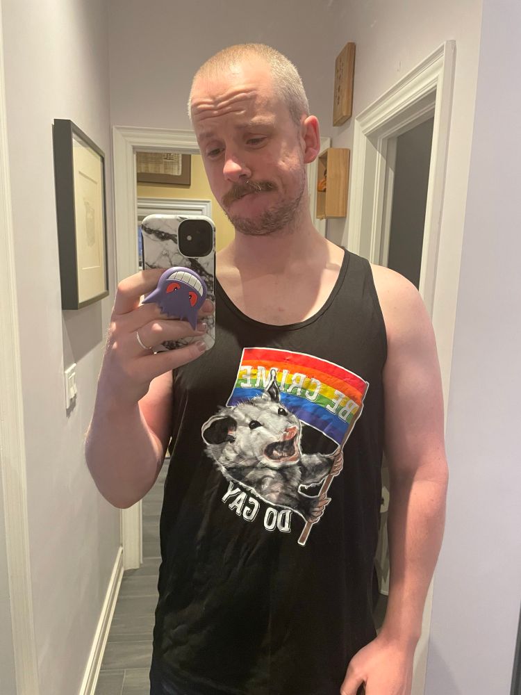 A white mustachioed man wearing a blank tank top that has an opossum waving a raibow flag that says "Be crime do gay" on it. He has a popsocket on his phone that is a Gengar and the gengar is upside down.