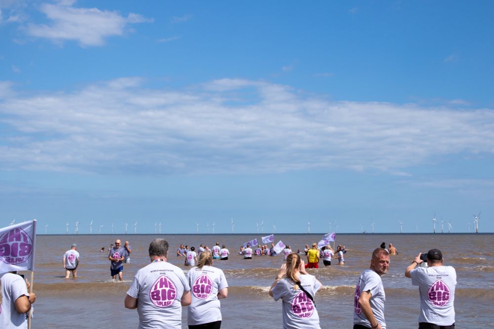 A group of people in the sea holding Big Splash flags, with others stood on the sand looking out to sea.