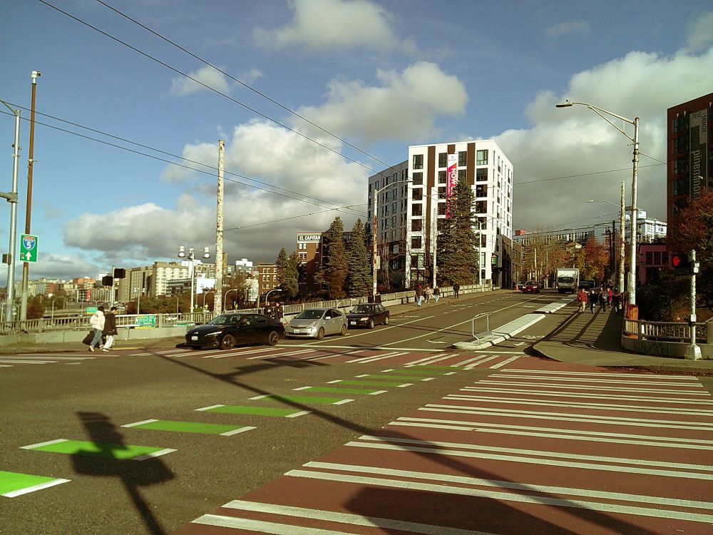 A large intersection, with a partly cloudy sky, long shadows of street signs, and people, cars, and buildings more distantly in the background.