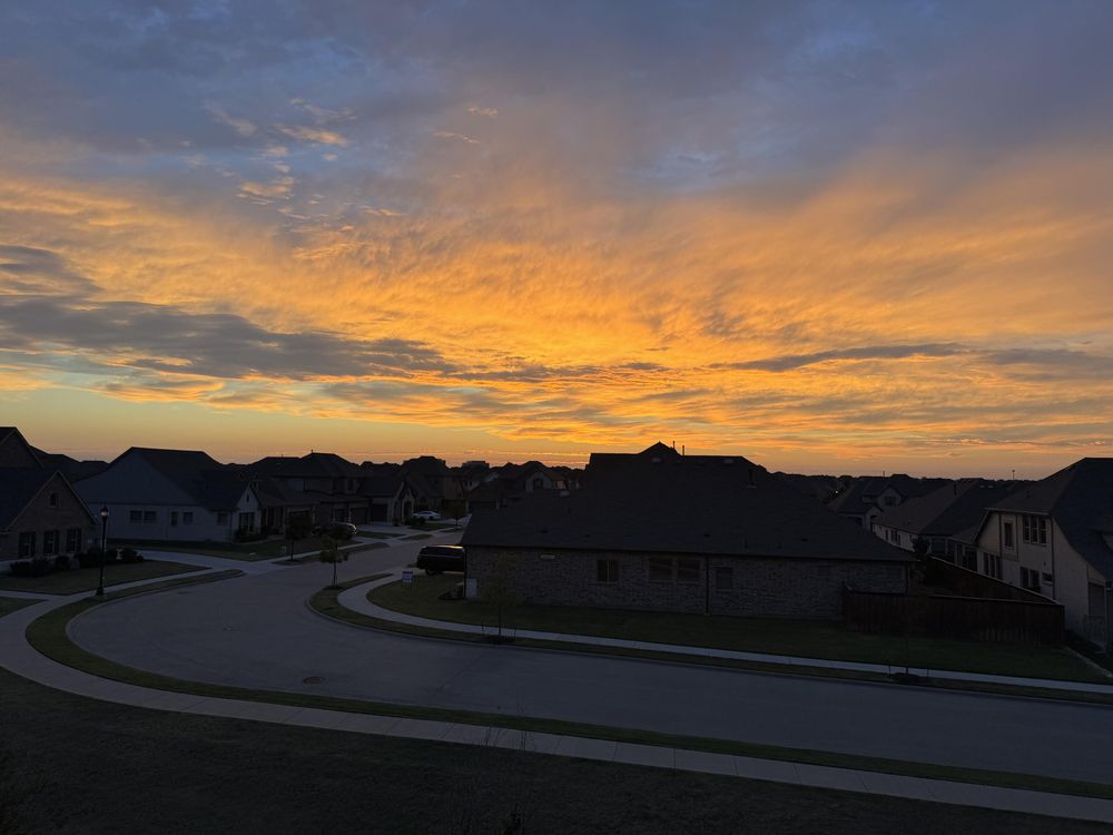 An elevated view of a quiet suburban neighborhood at sunrise or sunset. The sky is filled with a brilliant display of orange and yellow clouds, while the houses and a curving street below are cast in shadow.