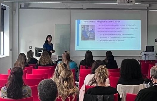 A small audience, seated on red chairs, is facing a person who is giving a presentation 