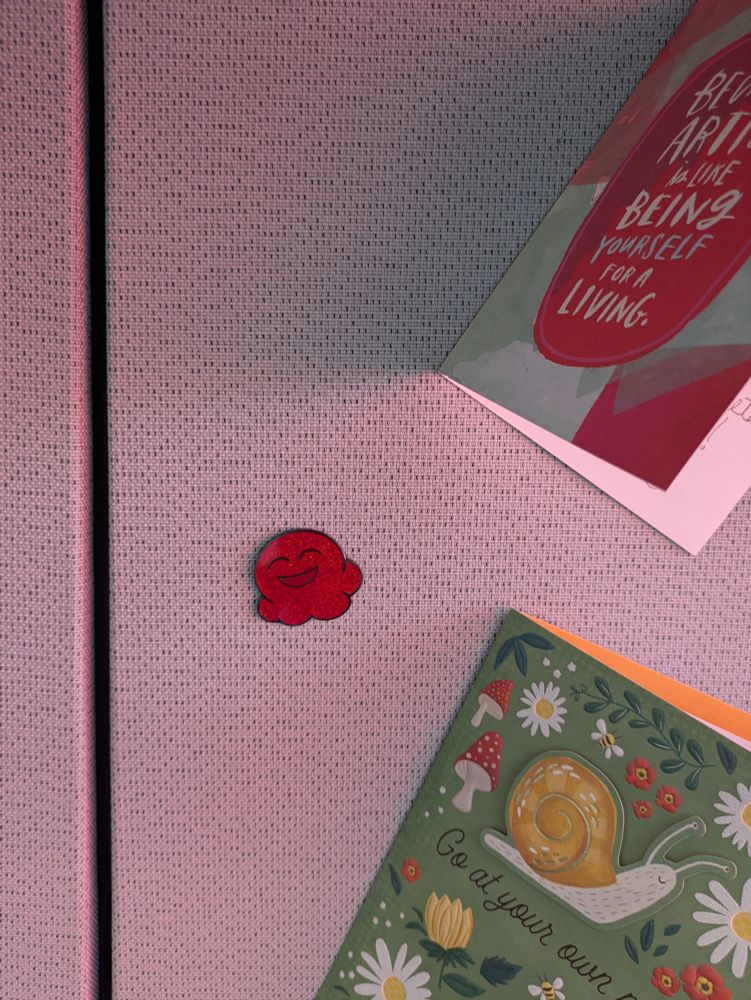 Small, red, smiling blob-shaped (reminiscent of an ice cream scoop) enamel pin on cubicle wall