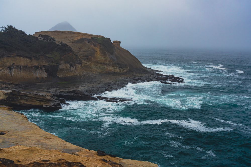 A dramatic view of Cape Kiwanda on the Oregon coast, where rugged sandstone cliffs meet the restless Pacific Ocean. The cliffs are layered in warm hues of golden yellow, tan, and deep brown, revealing intricate striations and grooves etched by centuries of wind, rain, and waves. The foreground features weathered sandstone, its surface a mix of smooth curves and sharp ridges, highlighting the ongoing interplay of erosion and resilience. Near the base of the cliffs, darker basalt formations emerge, their jagged edges standing firm against the powerful waves that crash and churn below. White foam froths against the rocks, sending sprays of water into the air, while swirling teal and deep blue waters create a dynamic contrast with the earthy tones of the land.

In the background, partially veiled by coastal mist, Chief Kiawanda Rock rises dramatically from the ocean. This towering basalt seastack, with its triangular silhouette, contrasts sharply with the softer, sculpted sandstone cliffs of the cape. It serves a vital role in the landscape, acting as a natural shield that absorbs much of the Pacific’s force, allowing the softer sandstone headland to endure despite its fragility. This unique interplay between the durability of basalt and the erodibility of sandstone has shaped Cape Kiwanda’s iconic appearance over time.

The misty, overcast sky adds to the scene’s moody, atmospheric quality, blending seamlessly with the ocean horizon and softening the contrast between land, sea, and sky. The light is diffuse, casting a muted glow that enhances the rich textures of the sandstone and the movement of the ocean. Small patches of greenery cling to the cliffs, their vibrant hues standing out against the weathered rock, a reminder of the tenacity of life even in harsh coastal conditions.

Cape Kiwanda is a dynamic, evolving landscape, a testament to the relentless power of nature and the surprising balance of strength and fragility.