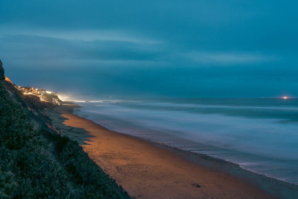 This long-exposure photograph captures a view of the Lincoln City, Oregon beachfront at dusk. The scene is dominated by a wide expanse of smooth, silky ocean waves, blurred from the long exposure to create a glass-like surface stretching to the distant horizon. The water, bathed in soft blue and teal tones, reflects the cool hues of the fading evening light under a thick, cloud-filled sky that adds an air of quiet melancholy. 

A strip of wet sand along the shoreline glistens faintly, catching the warm, golden glow of lights spilling from the cliffside homes above. These homes form a string of cozy, illuminated structures perched along the rugged cliff, with their soft yellow lights contrasting against the cool blues of the ocean and sky. The shoreline curves gently into the distance, creating a sense of depth and leading the eye toward faint, glowing points of light on the far horizon—likely offshore buoys or distant ships.

In the foreground, coastal vegetation blankets the steep slope of the cliff, appearing dark and textured in the low light. The sand below features a faint, circular impression, possibly from a previous visitor to the beach, adding a subtle touch of human presence to the otherwise tranquil and natural scene. The overall composition balances the warmth of artificial light against the cool, natural tones of the landscape, evoking a sense of peaceful solitude unique to the Oregon coast at dusk.