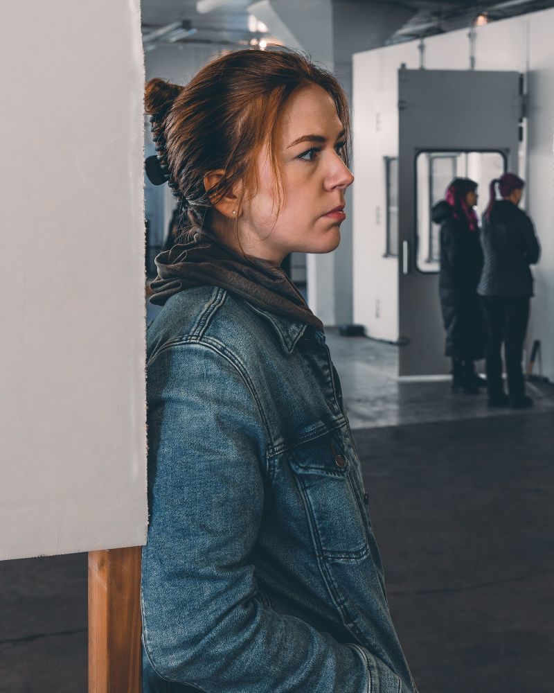 A woman with her red hair tied up, wearing a denim jacket, leans against a white wall, gazing intently into the distance. Her expression is thoughtful, as though lost in deep contemplation. 

In the background, two people with pink hair are seen conversing, slightly blurred, creating a sense of focus on the woman in the foreground. The industrial-like setting adds to the introspective mood of the scene.