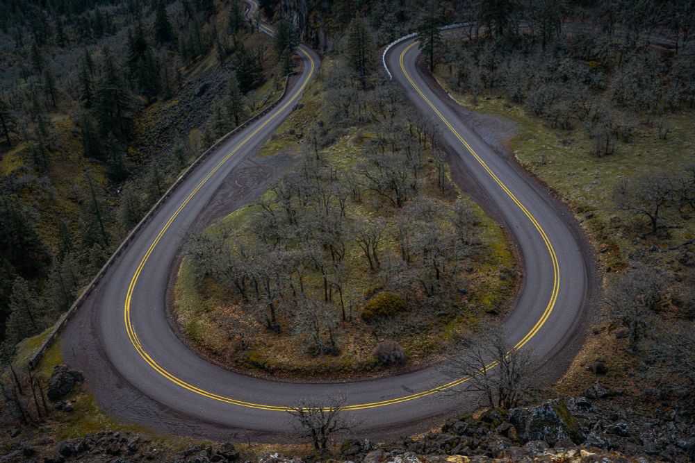 Elevated shot of a serpentine bend along the Historic Columbia River Highway at Rowena Crest, contrasting with the landscape of sparsely wooded areas and rocky terrain.