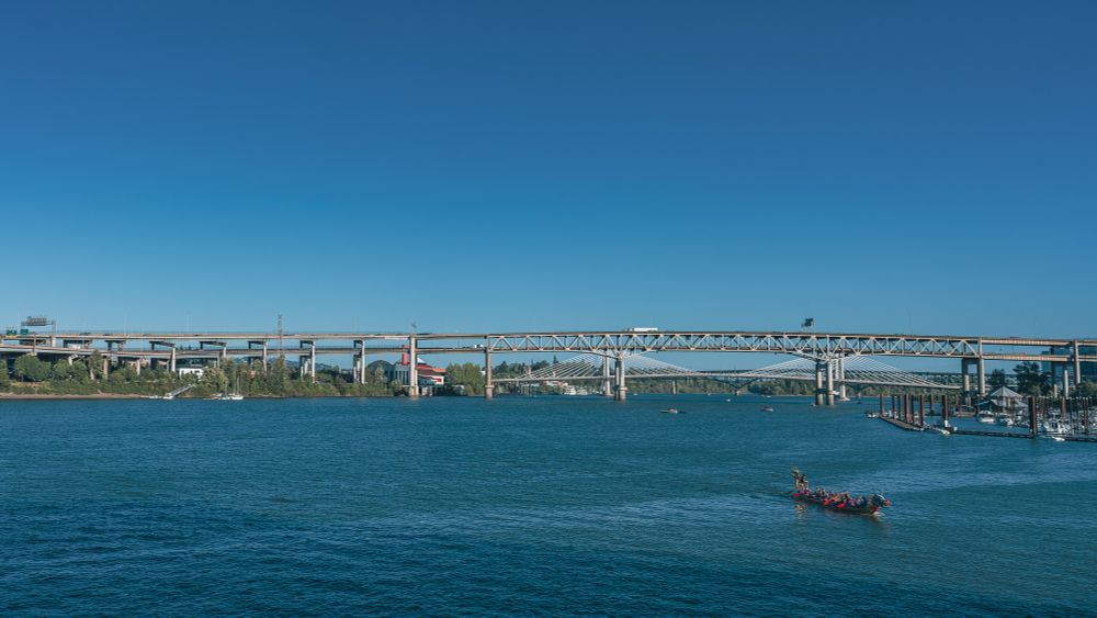 A vibrant dragon boat glides across the blue waters of the Willamette River in Portland, Oregon, under the expansive Marquam Bridge. The boat is rowed by a team of paddlers, and the stern is adorned with a decorative dragon head. 

In the background, the distinctive bridge, known for being Portland's busiest, stretches across the river. On the right (west) bank, a marina is visible. On the left (east) bank you can see the Oregon Museum of Science and Industry (OMSI) and the decommissioned submarine USS Blueback, which appeared in the film "The Hunt for Red October." 

The clear blue sky adds to the crisp and lively atmosphere of the scene.