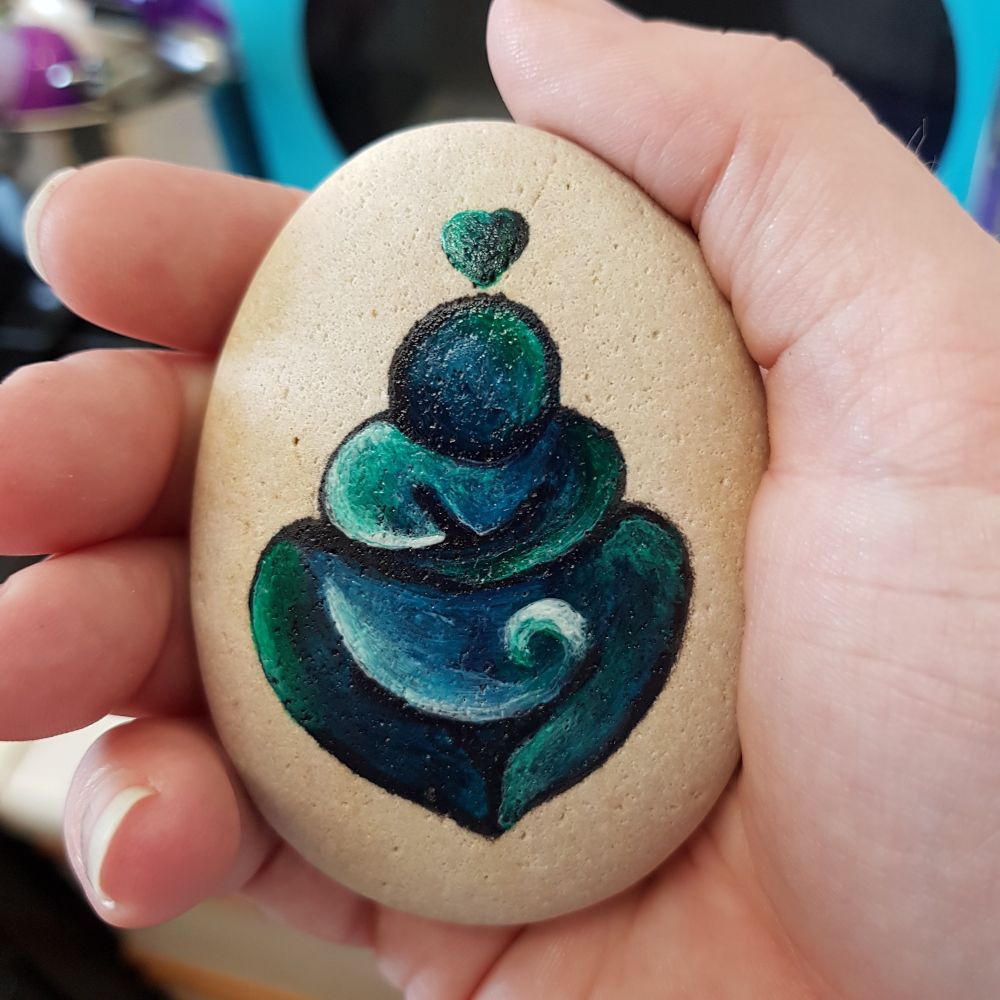 A hand gently clasps a pale beige ovoid pebble. It's roughly palm sized. On it is a simplistic drawing of a human figure. There are few features; a round head atop a torso that is merely a pair of arms that come to points. These arms are wrapped around the torso, as if the figure is hugging itself. Below that and framed by enormous thighs (that likewise come to points) is an equally large belly upon which a white crested wave is depicted. The whole figure is painted in shades of sea-greens and blues. Surmounting the figure is a small heart.