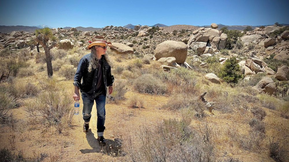 Portrait of yours truly walking through the desert just outside Joshua Tree, near Pioneertown. Mexican palm leaf straw cowboy hat, jeans, black t-shirt, striped pearl snap overshirt, if you must know the details. Bonus for alt text readers: Pioneertown was built as a location set for Hollywood westerns around the 40's or 50's (I think). Abandoned after the filming dried up, it's been since reoccupied by private residences and  local shop owners, and it still remains its TV Western charm. 