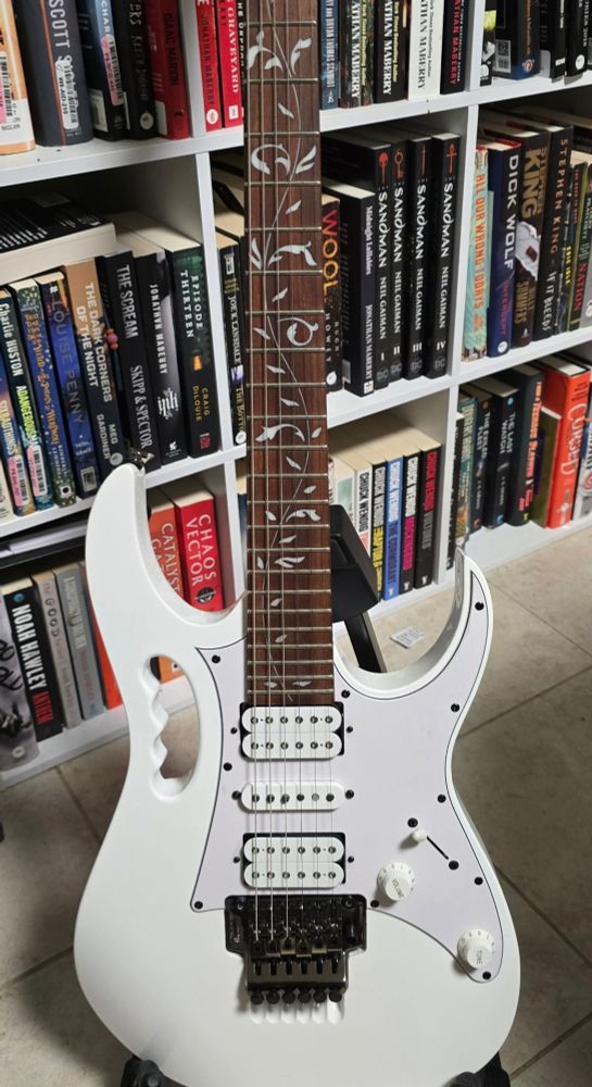 A white guitar, Steve Vai signature, with a vine inlay on the fretboard and a "monkey grip" handle on the side.