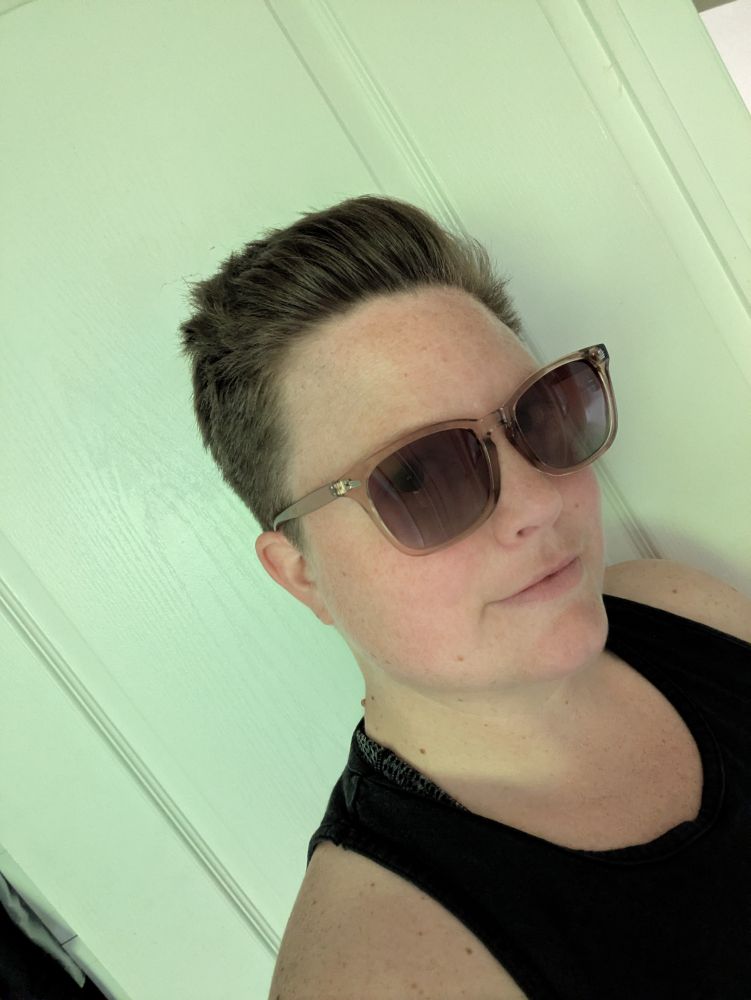 A white person with short hair poses in a black tank top and sunglasses. 