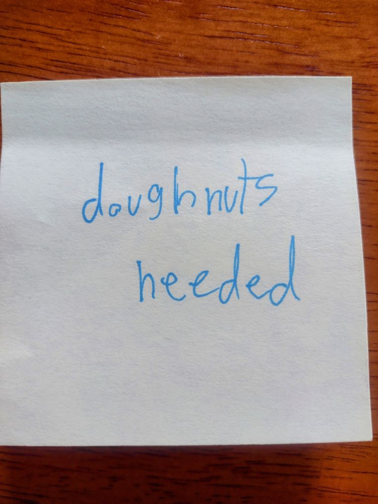 A yellow post it on a wooden table. The post it is kid handwriting in blue ink and says "doughnuts needed"