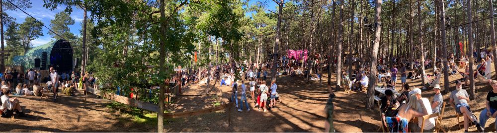 Een panorama van het podium op de open plek. Mensen tussen de dennenbomen.