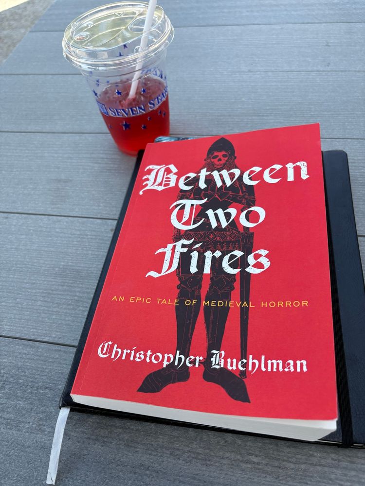 A book with a red cover sits on top of a black notebook. The book is titled Between Two Fires by Christopher Buehlman. The notebook and book are on top of a grey table. There’s a clear cup of iced hibiscus tea behind the stack.