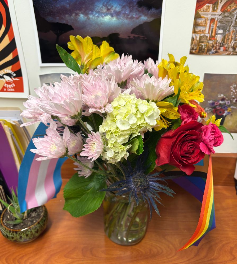 a mason jar of flowers of all colors. a trans flag and progress pride flag are also in the jar. The jar is sitting on a brown desktop with photos taped to the wall in the background.
