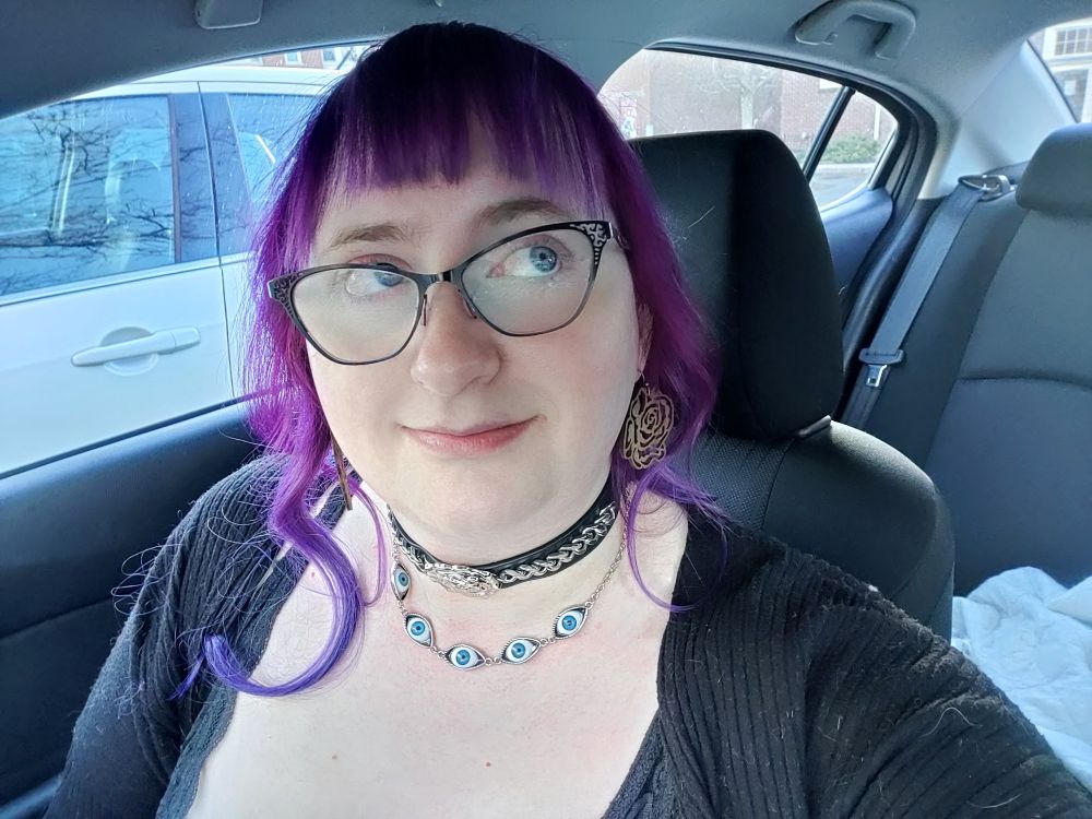 Runa is sitting in her car, wearing a low cut black lace dress, lace gloves, and a choker detailed with chain and a metallic rose. Her hair is freshly dyed a bright, warm purple fading into a cooler, darker purple at the ends. Hey hair is about three feet long and curled slightly into loose waves and she is wearing blunt bangs. In this image she is smiling and looking toward to her right.