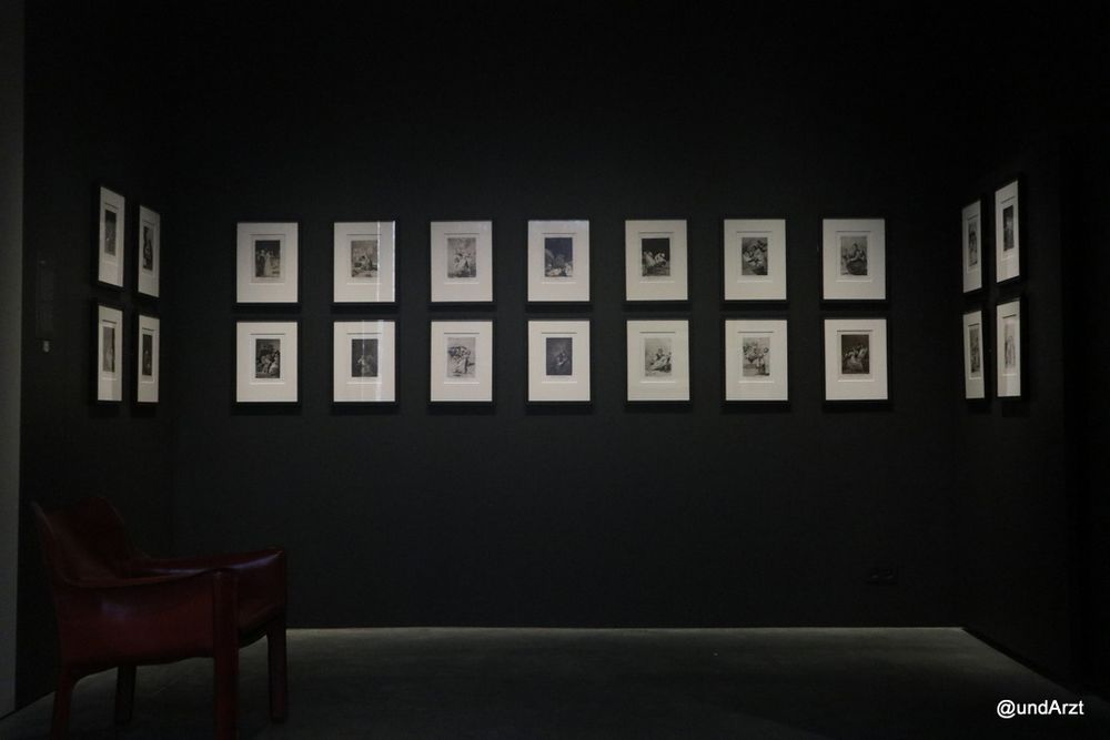 Blick auf eine dunkle Wand, an der 14 gleich große Bilder hängen, rechts und links jeweils noch 4 gleichartige. Links steht ein dunkler Sessel auf leicht hellerem Boden.