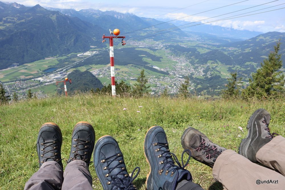 Sechs Wanderschuhe vor Alpenpanorama mit Versorgungsseilbahn über rot-weißen Trägern