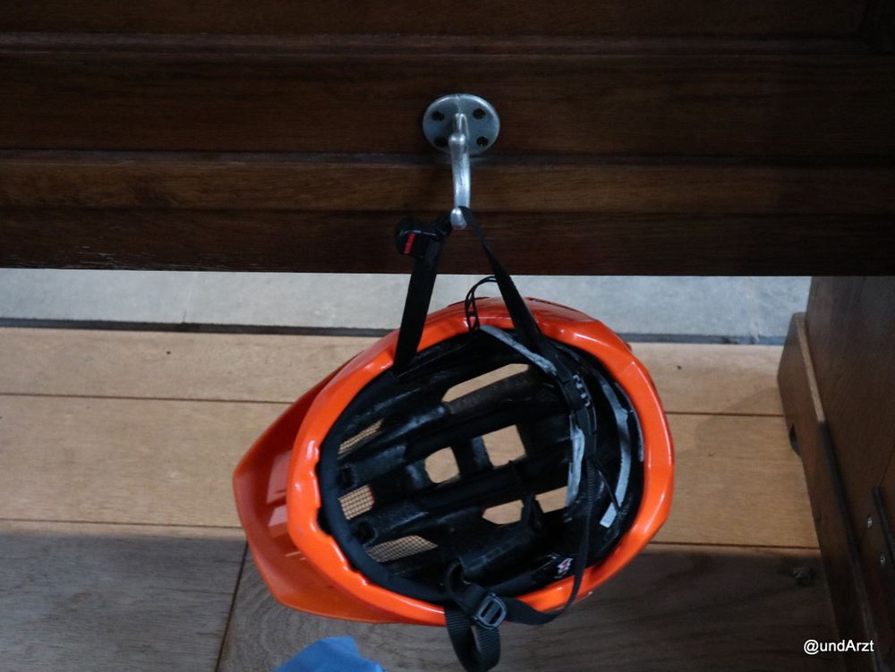 Mein orangefarbener Fahrrad-Helm bei einem Konzert hängt an einem Haken in der Kirchenbank.