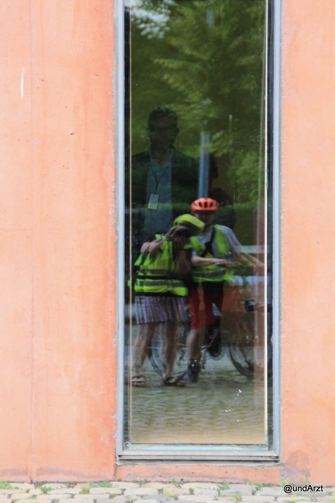 In einem hohen Fenster spiegeln sich zwei Radfahrer und ein Baum. hinter dem Fester erspäht man eine Silhouette eines Manns in einem Jackett mit weißem Hemd.