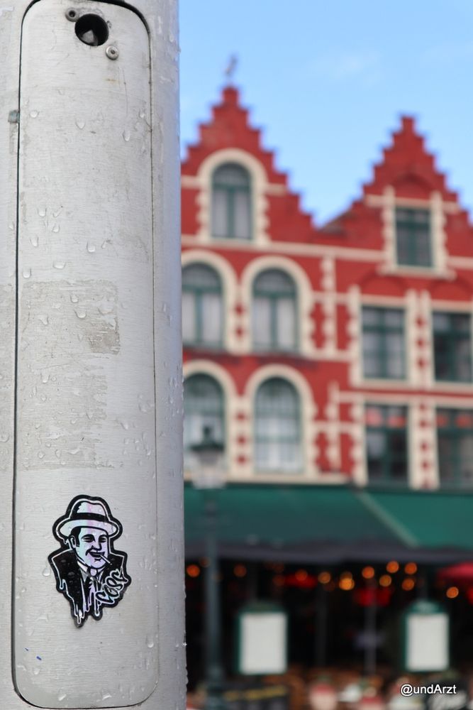 Ein schwarz-weißer Aufkleber auf einem Laternenmast, zeigt einen Mann mittleren Alters mit Hut und Zigarette (ich assoziiere das mit Mafia-Filmen). Im Hintergrund rechts, unscharf, alte Häuser aus roten und weißen Ziegeln mit Restauration unter grünen Vordächern.