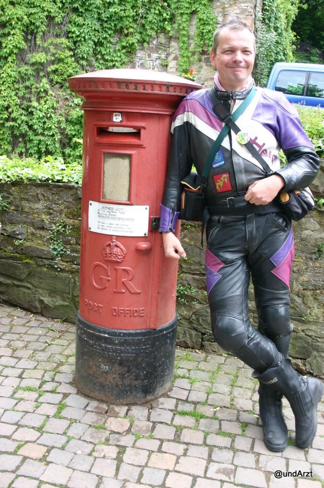 roter Briefkasten in Tonnenform (GR post office), daran lehnt ein junger (nun ja, 45-jähriger) Mann in schwarz-weiß-violetter Harro-Lederkombi und feistem Grinsen.