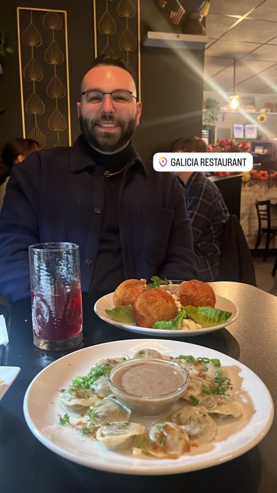 A photo of Gianluca smiling at Galicia Restaurant in Rochester, NY. He’s sitting at a table with banosh, varenyky, and a glass of kompot.