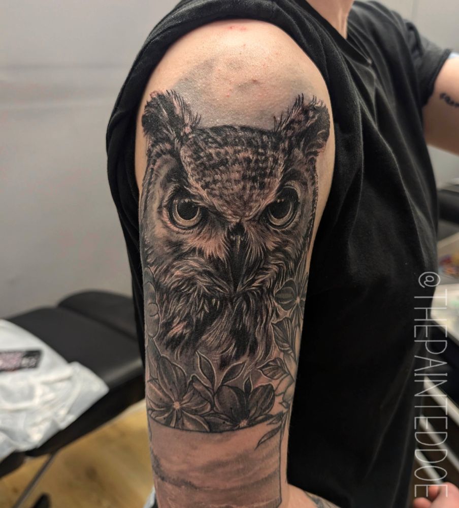 An after photograph of a fresh owl tattoo on someone's upper arm, covering the owl in the photograph previous. It is tattooed from the neck up in black and grey, with it's eyes looking directly at the camera.