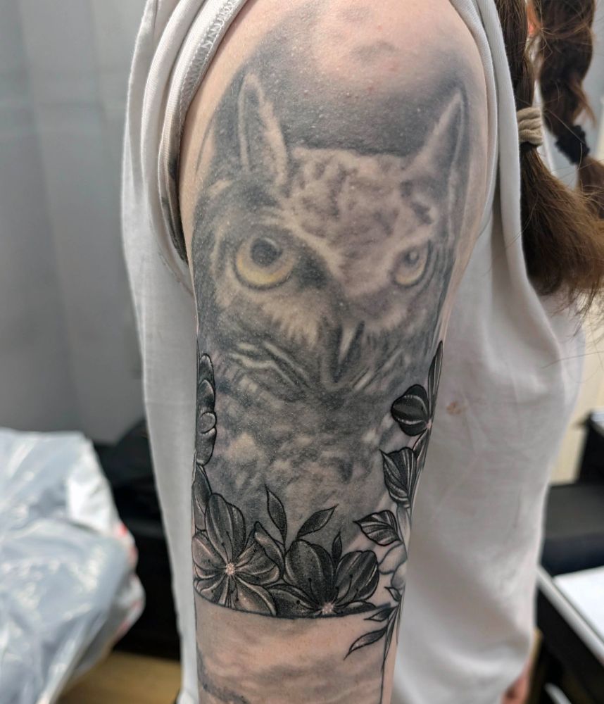 A before photograph of an old owl tattoo on an upper arm. The face is very faded and out of proportion.