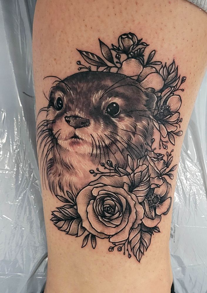 A photograph of an illustrated tattoo of an otter portrait surrounded by flowers and foliage, the tattoo is in black and grey.