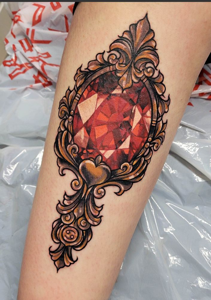 A photograph of a large leg tattoo, it shows a large ornate hand frame in copper/gold with a large red ruby stone set into the centre where a mirror would be. There is a small engraving of the cancer star sign set into the lower part of the handle and a heart shape featured into the upper part of the handle.